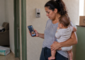 Mulher segura um bebê no colo e observa no celular a fotografia de uma encomenda deixada diante de outra porta, com uma caixa de entrega ao fundo.