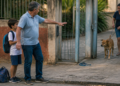 Homem idoso estende o braço de forma protetora diante de um menino com mochila na calçada, enquanto uma mulher tenta conter um cão próximo ao portão aberto de uma casa.