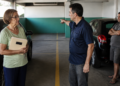 Mulher idosa com pasta nas mãos conversa com um homem que aponta para vagas de garagem em um estacionamento, enquanto outro homem observa ao lado de um carro.