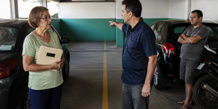 Mulher idosa com pasta nas mãos conversa com um homem que aponta para vagas de garagem em um estacionamento, enquanto outro homem observa ao lado de um carro.