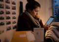 Mulher em casa à noite observa o celular com expressão preocupada, ao lado de uma caixa e de documentos, diante de uma parede com várias fotos de carros.