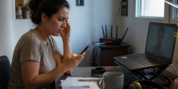 Mulher sentada em uma mesa de trabalho olha para o celular com expressão de preocupação, diante de um notebook ligado e de um roteador com luzes acesas ao fundo.