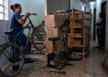 Mulher conduz uma bicicleta por um corredor de prédio com caixas, estante e objetos acumulados, enquanto outra moradora observa ao fundo.