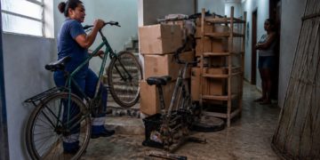 Mulher conduz uma bicicleta por um corredor de prédio com caixas, estante e objetos acumulados, enquanto outra moradora observa ao fundo.