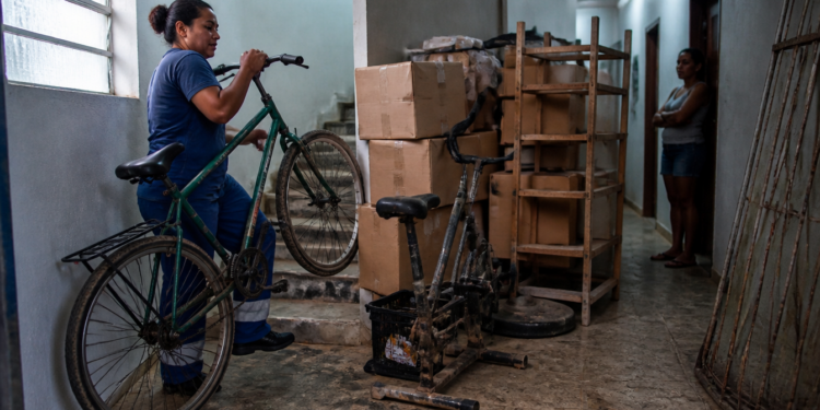 Mulher conduz uma bicicleta por um corredor de prédio com caixas, estante e objetos acumulados, enquanto outra moradora observa ao fundo.