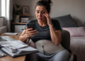 Mulher com faixa abdominal consulta o celular diante de um notebook, documentos e medicamentos, enquanto uma muleta permanece ao lado da cama.