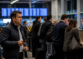 Homem com celular e mala de viagem aguarda diante de uma fila de passageiros em um balcão de atendimento de aeroporto, com painéis de voos desfocados ao fundo.