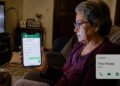 Mulher idosa sentada no sofá segura o celular com uma conversa aberta, enquanto observa a tela com expressão preocupada, em uma sala de casa com televisão ligada e uma xícara sobre a mesa.