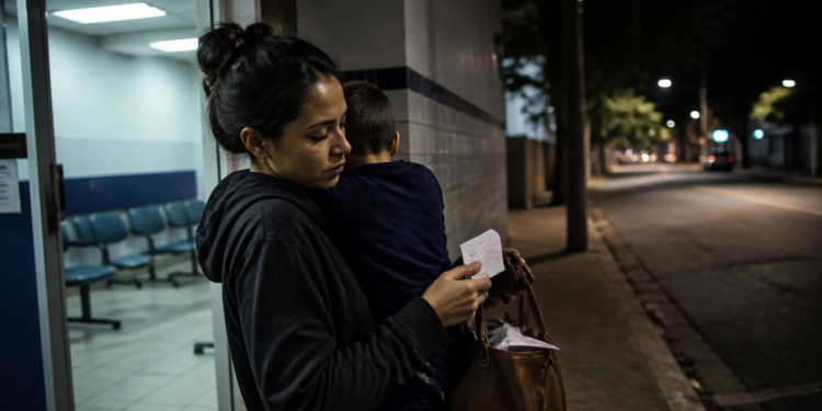 Mulher segura uma criança no colo e confere um pequeno papel diante da entrada iluminada de uma unidade de atendimento durante a noite.
