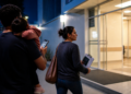 Mulher caminha em direção à entrada iluminada de uma unidade de atendimento enquanto segura papéis, acompanhada por um homem com uma criança no colo e um celular na mão.