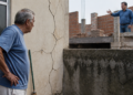 Aposentado observa grandes rachaduras na parede externa de sua casa enquanto o vizinho gesticula em uma obra de alvenaria ao lado.