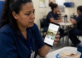 Mulher com uniforme azul segura um celular que exibe a foto de uma criança, enquanto está sentada diante de uma refeição em um ambiente de trabalho.