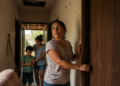 Mulher fecha a porta de um quarto enquanto observa insetos próximos ao teto, com outra mulher e uma criança ao fundo do corredor.