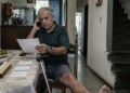 Homem idoso fala ao telefone enquanto lê um documento sentado à mesa de casa, com papéis e uma bengala ao lado.