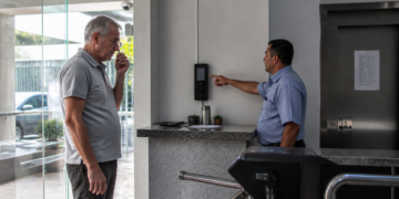 Homem idoso observa enquanto funcionário aponta para um aparelho de controle de acesso instalado na parede da portaria de um condomínio.