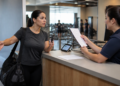 Mulher com bolsa esportiva conversa de forma tensa com uma atendente no balcão de uma academia, enquanto segura o celular e a funcionária analisa um documento.