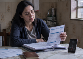 Mulher sentada à mesa da cozinha analisa uma conta impressa ao lado de uma pasta com documentos, outros boletos e um celular com chamada em andamento.