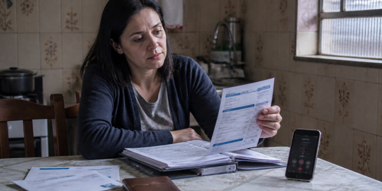 Mulher sentada à mesa da cozinha analisa uma conta impressa ao lado de uma pasta com documentos, outros boletos e um celular com chamada em andamento.