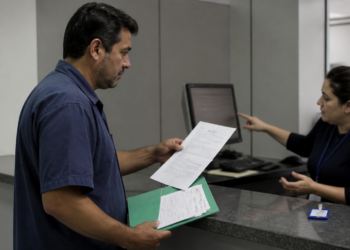 Homem segura documentos e uma pasta verde em um balcão de atendimento enquanto uma funcionária aponta para a tela do computador em um ambiente administrativo.