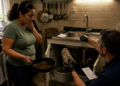 Mulher segura uma frigideira com líquido escuro na cozinha enquanto um profissional mostra resíduos retirados do encanamento sob a pia.