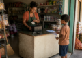 Mulher atrás do balcão de uma pequena mercearia segura um papel e mexe no caixa, enquanto um menino em pé à frente também segura papéis.
