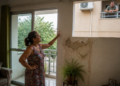 Mulher aponta para uma grande mancha de infiltração na parede de seu apartamento enquanto um vizinho observa pela janela do prédio ao lado.