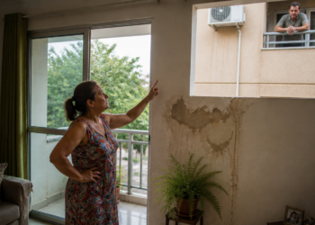 Mulher aponta para uma grande mancha de infiltração na parede de seu apartamento enquanto um vizinho observa pela janela do prédio ao lado.