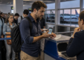 Passageiro com mochila apresenta o celular e um comprovante de viagem a uma atendente no balcão do aeroporto, enquanto outras pessoas aguardam na fila com suas bagagens.