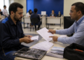 Técnico de manutenção segura comprovantes enquanto conversa com um atendente que aponta para um formulário em uma agência, com um computador desmontado sobre a mesa.