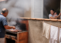 Homem prepara carne em uma churrasqueira no quintal, com muita fumaça subindo perto do muro, enquanto uma mulher com um bebê observa pela janela da casa ao lado.