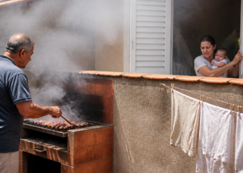 Homem prepara carne em uma churrasqueira no quintal, com muita fumaça subindo perto do muro, enquanto uma mulher com um bebê observa pela janela da casa ao lado.
