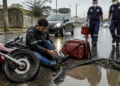 Entregador sentado no asfalto molhado ao lado de uma motocicleta caída e de um grande buraco com água, enquanto dois socorristas do SAMU se aproximam com equipamentos de atendimento.